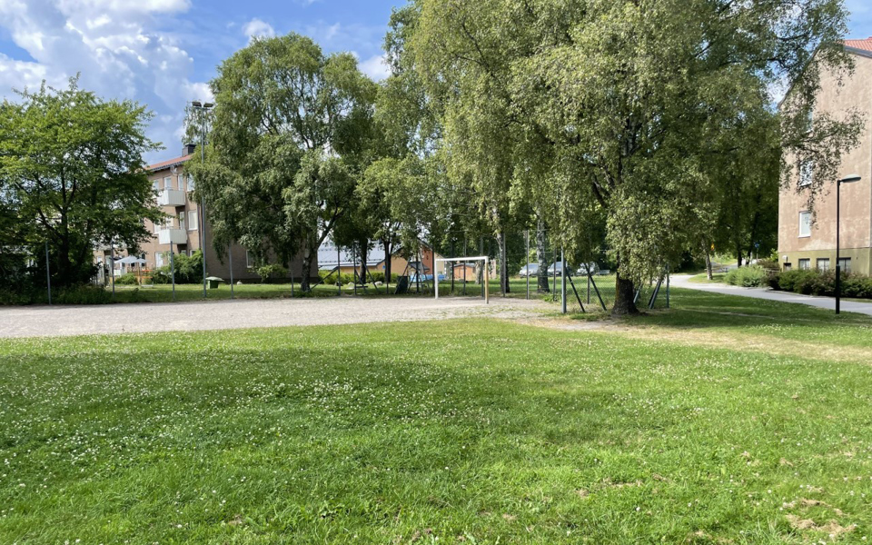 En grusbollplan med fotbollsplan intill en gräsmatta. Bolplanen är delvis inhägnad och bakom planen ses höga träd och flerbostadshus. Foto